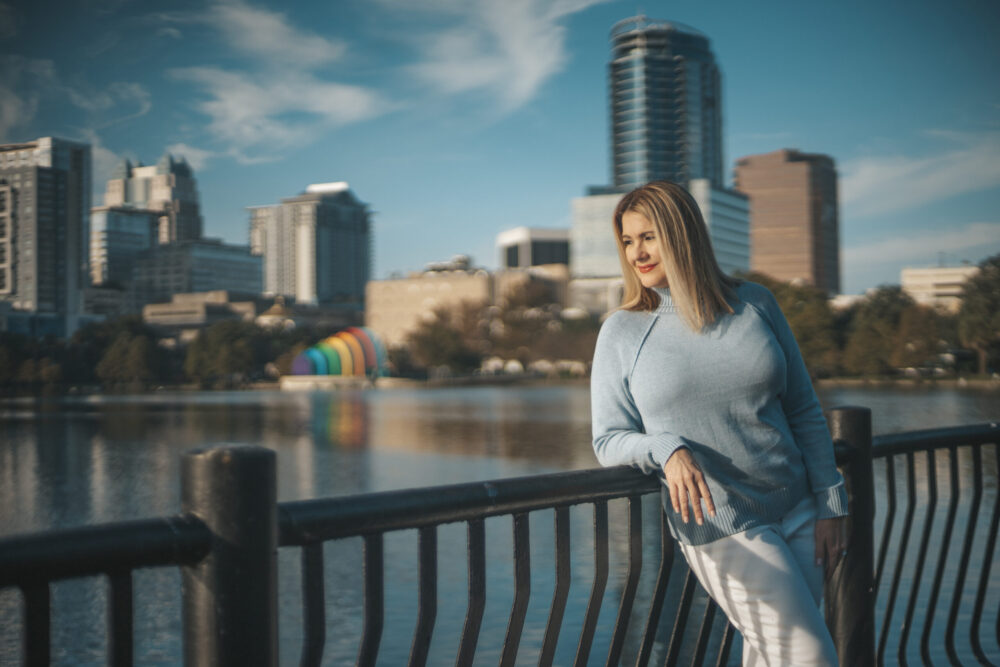 Picture of Jill Carlyle standing in front of a railing. There is water and tall buildings behind her. Jill is a Reinvention Rebels who has reinvented herself in her 40s and 50s.