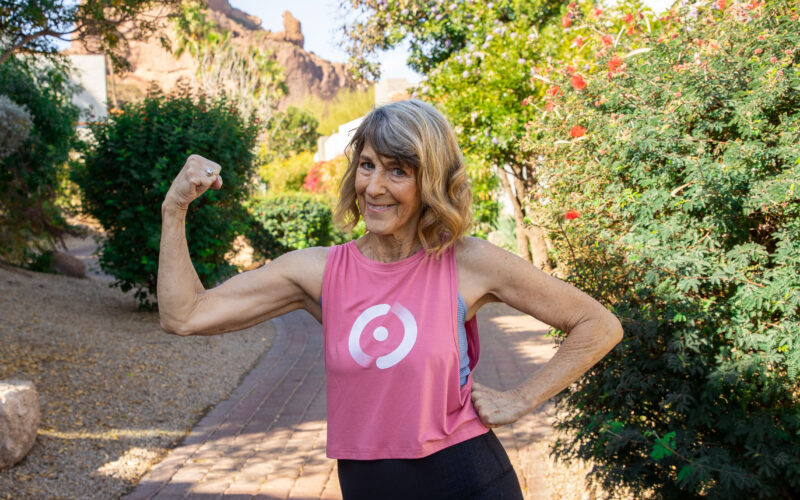 Picture of Reinvention Rebel Vickie Aigner. She's wearing a pink sleeveless workout top and flexing her biceps. Behind her is an outdoor setting.