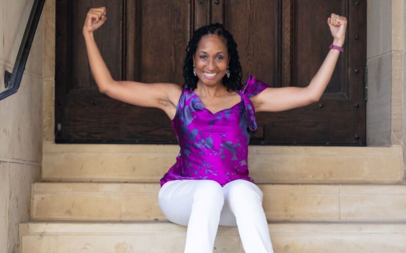 Picture of Wendy Battles sitting on the steps outside of a building. She is flexing both arms to make muscles. Her motto is reinvent yourself no matter your age, it's never too late.