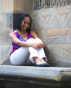 Midlife has taught Wendy Battles important lessons about slowing down and coming home to herself. She's pictured in a purple top and white pants sitting on a stone bench with her arms wrapped around her knees.