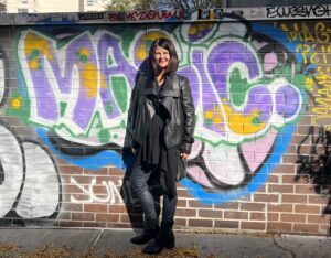 Midlife reinvention invites us to grow, be bold, have courage and create a life we love. Picture of Elizabeth in a black leather jacket standing in front of a brick wall with graffiti on it.