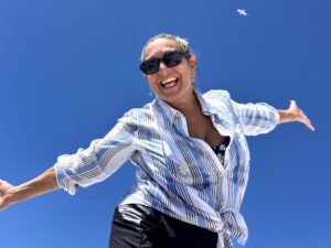 Friendship after 50 is possible for all of us. Meeting new people who help us become our best, most authentic selves. Picture of Stacey Tessa K Battat smiling with her arms wide open against a blue sky
